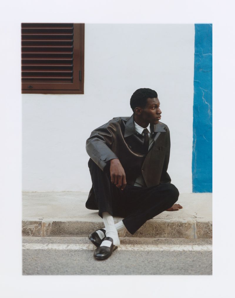 Portrait of a Black man in a brown jacket and black pants sitting on stone steps against a building with a window and blue accent stripe.