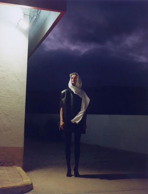 Fashionable adult stands beneath a light fixture outside against a dark, stormy sky, wearing a scarf over their head.