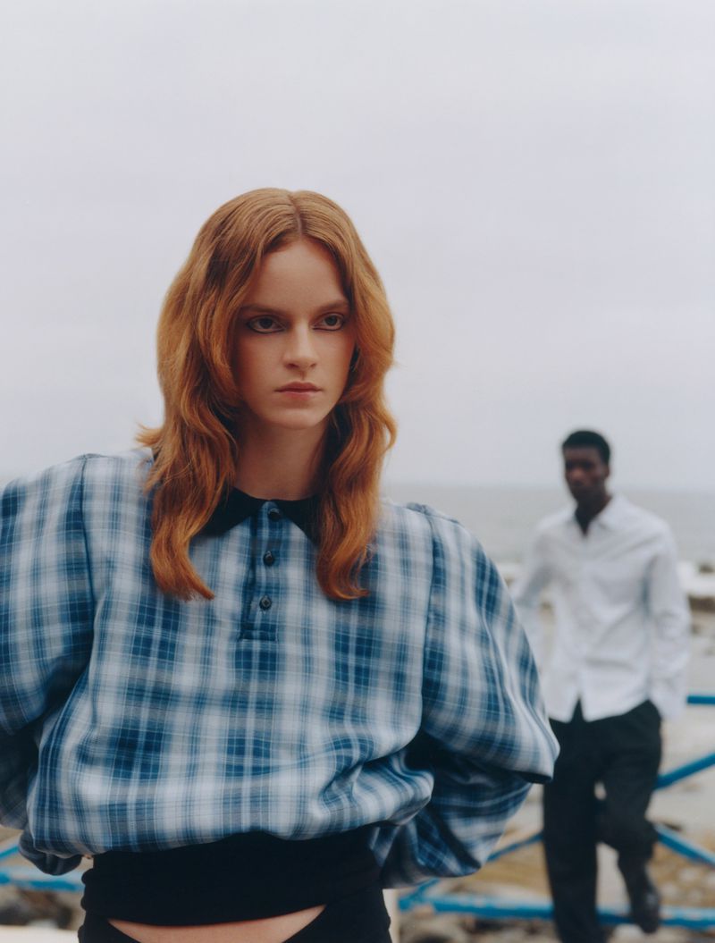 Two fashion models pose in an outdoor setting, woman with auburn hair in a blue plaid blouse, man with white shirt in the background.