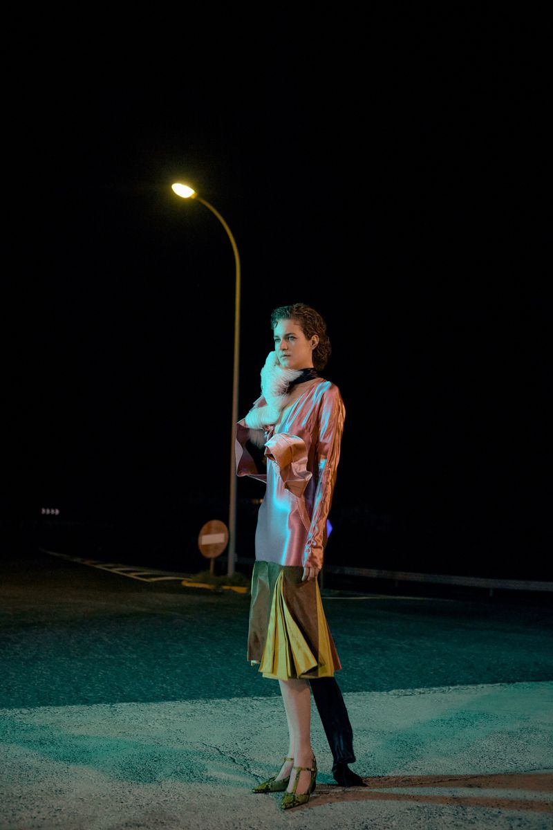 Elegant woman with fur stole posing at night near a street lamp in a stylish dress with green heels on an empty road.