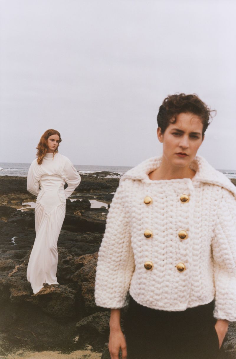 Two women in elegant fashion on a rocky beach under an overcast sky. One wears a white dress, the other a knit jacket and dark pants.