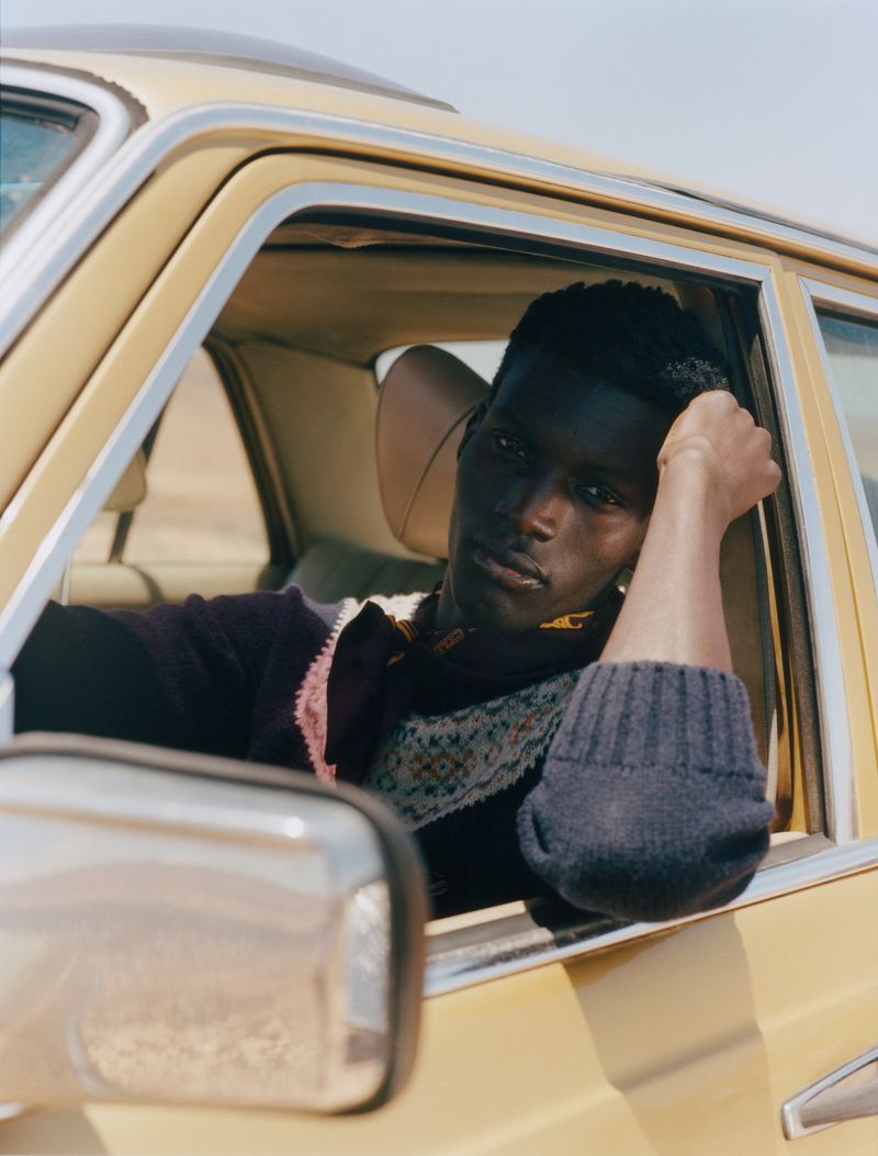 Portrait of a Black man inside a yellow car, gazing out the window with a relaxed expression, wearing patterned sweater, desert backdrop.