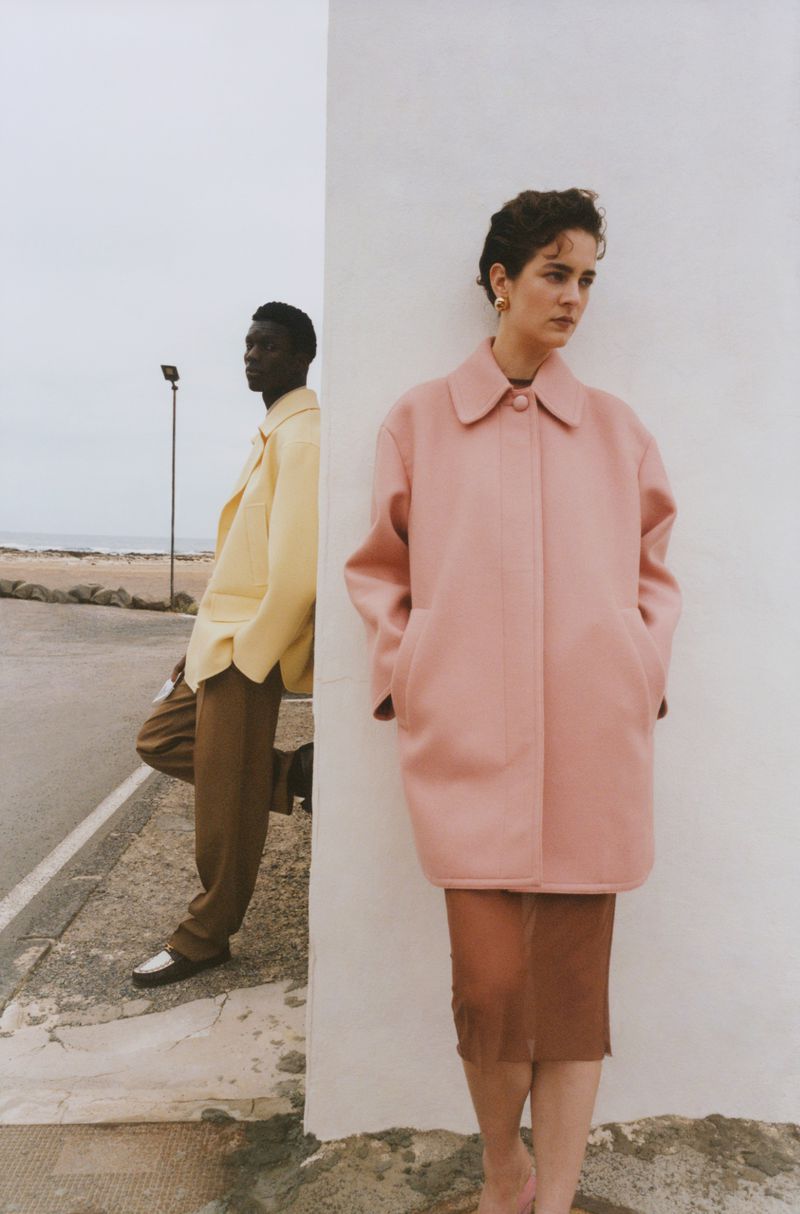 Two fashion models pose outdoors; an African American man in yellow jacket, a white woman in pink coat against a white wall.