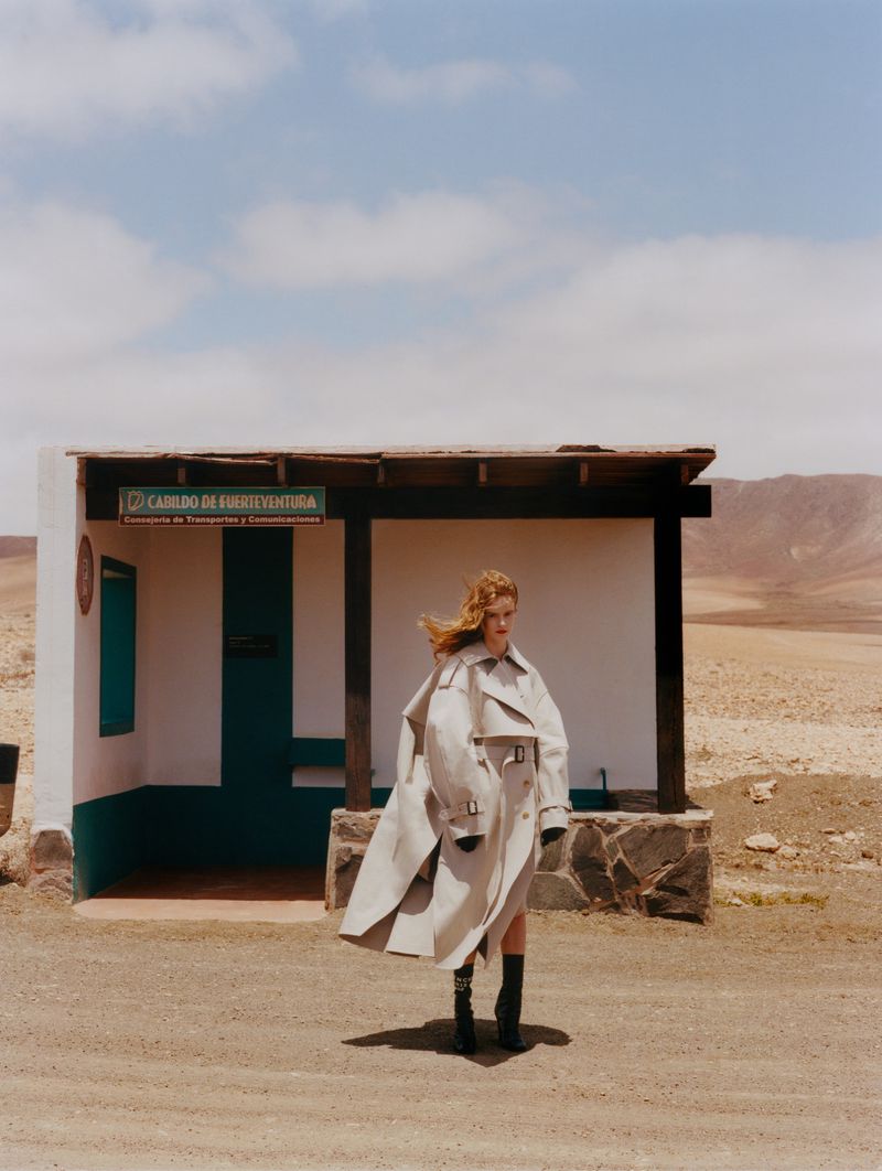 Adult woman poses in an oversized trench coat near a bus stop in a desert landscape with a clear blue sky during daytime.