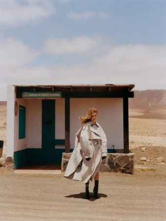 Adult woman poses in an oversized trench coat near a bus stop in a desert landscape with a clear blue sky during daytime.