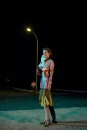 Model standing on a dark asphalt road at night under a street light, wearing a pink satin top and pleated color-blocked skirt.