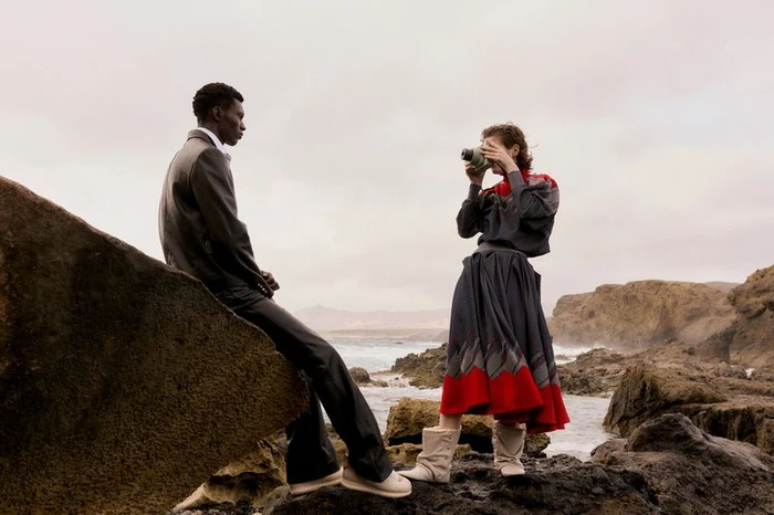 Model seated on rock looking at a woman photographing with a vintage camera on a rocky coast