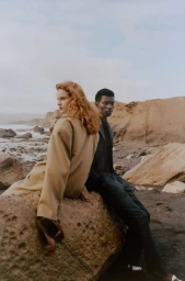 Two fashion models in tailored beige and dark coats resting on a boulder along a rocky, overcast coastal shoreline.