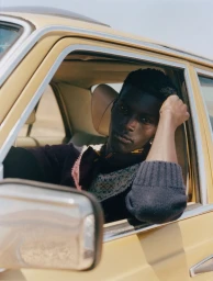 A man posing inside a vintage yellow car resting his arm on the open window frame for Mixte magazine.