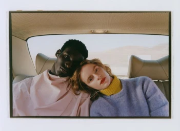 Two models seated inside a car sharing earphones, wearing a pink top and blue knit sweater against a distant desert landscape