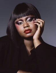 Model with dark bob hairstyle and dramatic eye makeup rests hand against cheek wearing a black shirt