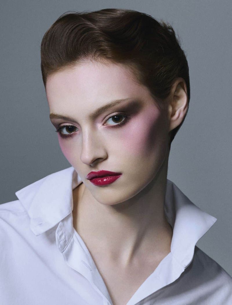 Close-up portrait of a redhead woman with heavy makeup, wearing a white collared shirt against a grey backdrop in a photo studio.
