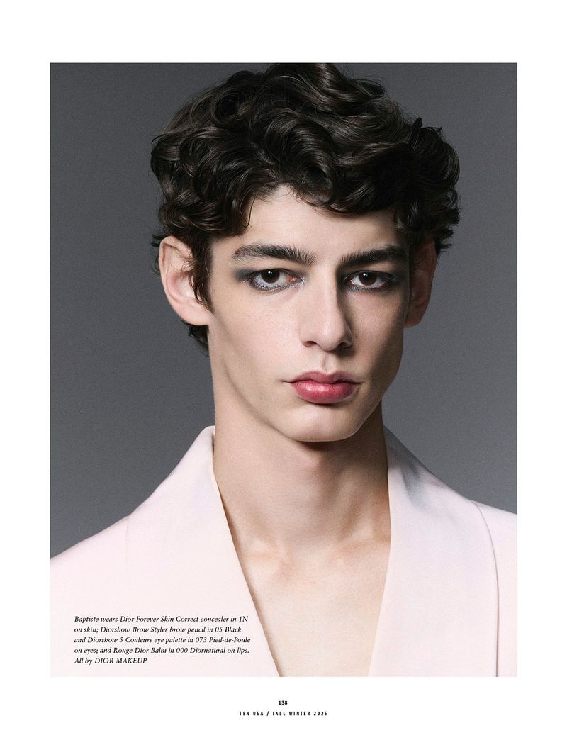 Close-up studio portrait of a young adult male fashion model with dark hair, wearing eye makeup and a white jacket, against a grey backdrop.