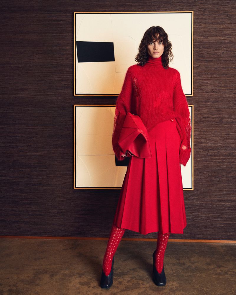 Fashionable woman posing in red sweater, skirt, patterned stockings, and dark heels in front of modern art and dark-brown wall.