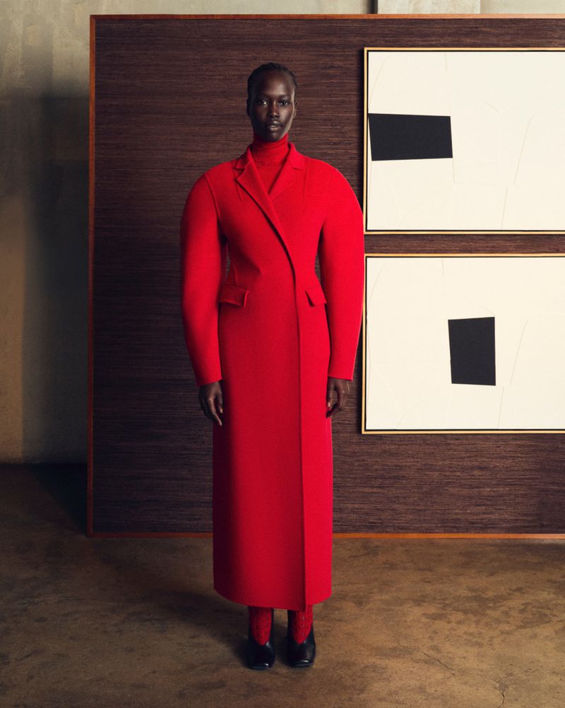 Elegant Black woman in a vibrant red coat, red leggings, and black heeled shoes poses in front of abstract art for high fashion portrait.