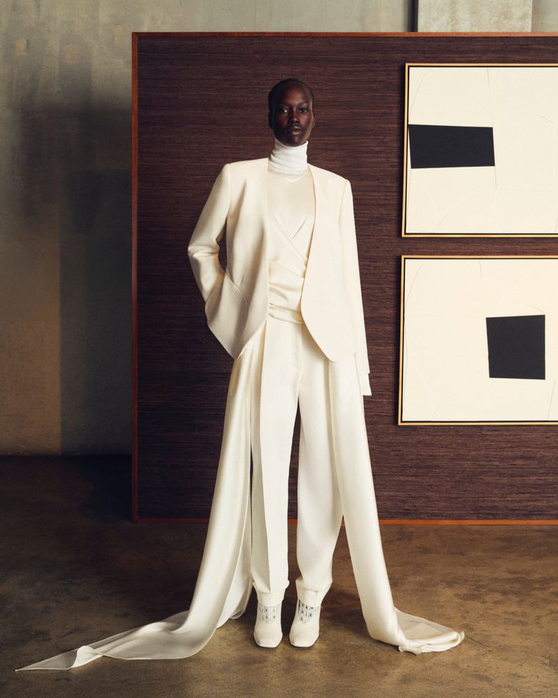 Elegant African-American woman in a white suit, with a white turtleneck and long white tails on the trousers, posing in a studio.