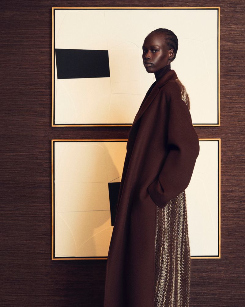 A tall, elegant Black woman in a long brown coat poses in front of framed abstract art against a dark brown patterned wall.