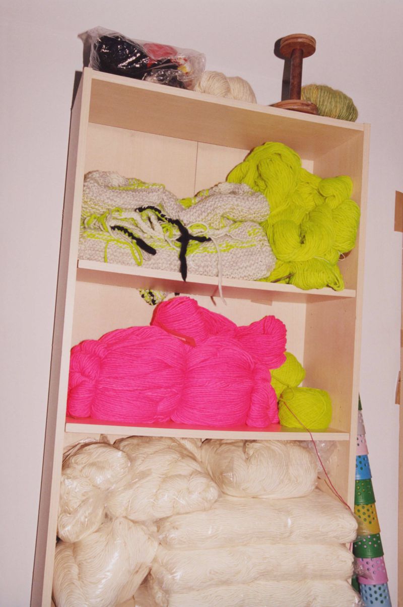 Bright neon pink, green, and white yarn on a wooden shelf. Textile supplies are stacked and organized in a crafting or hobby room.