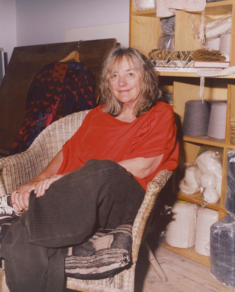 An adult woman sits in a wicker chair in a room, wearing a red shirt and brown pants, with shelves of yarn behind her.