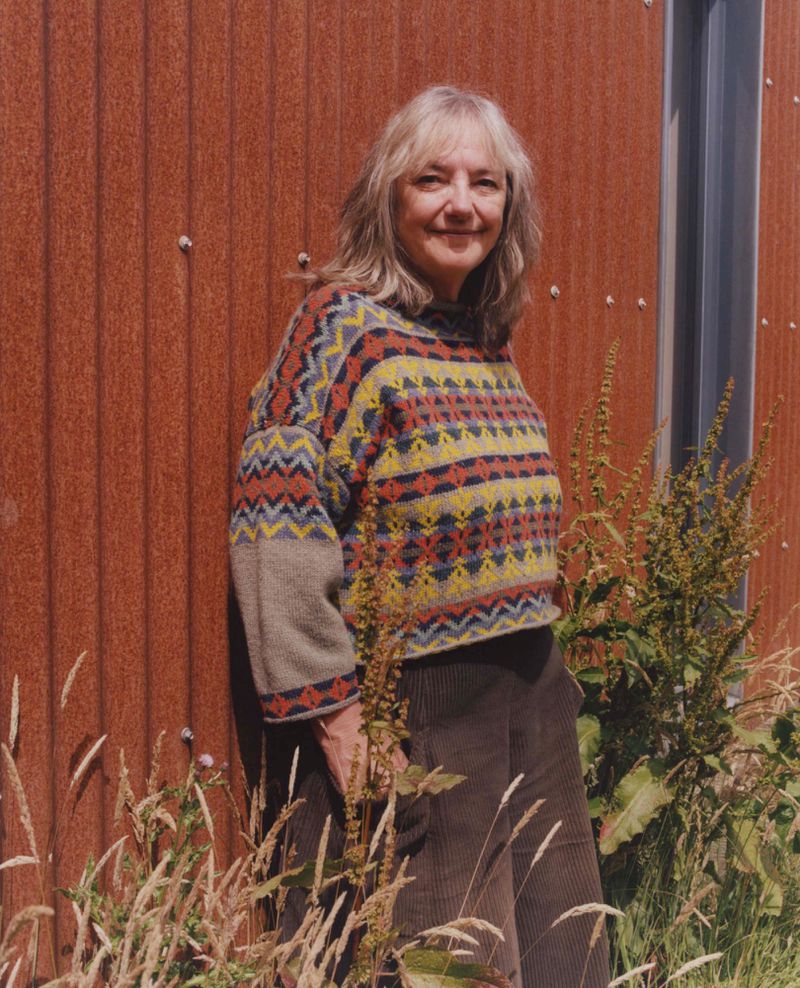 Mature woman with fair hair stands by a textured orange wall, wearing a colorful patterned sweater with green plants in foreground.