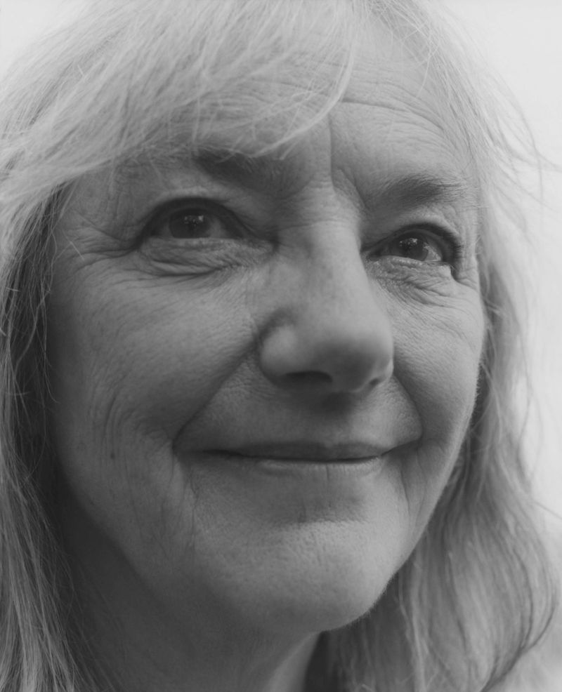 Monochrome portrait of a mature woman with long grey hair smiling gently at the camera in a close-up studio setting.