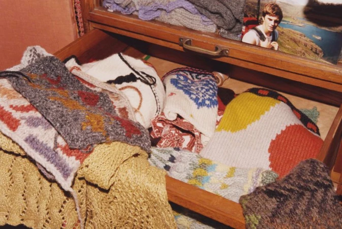 Open wooden drawer filled with colorful folded knitted garments, accompanied by a photograph of a young adult male in an indoor setting.
