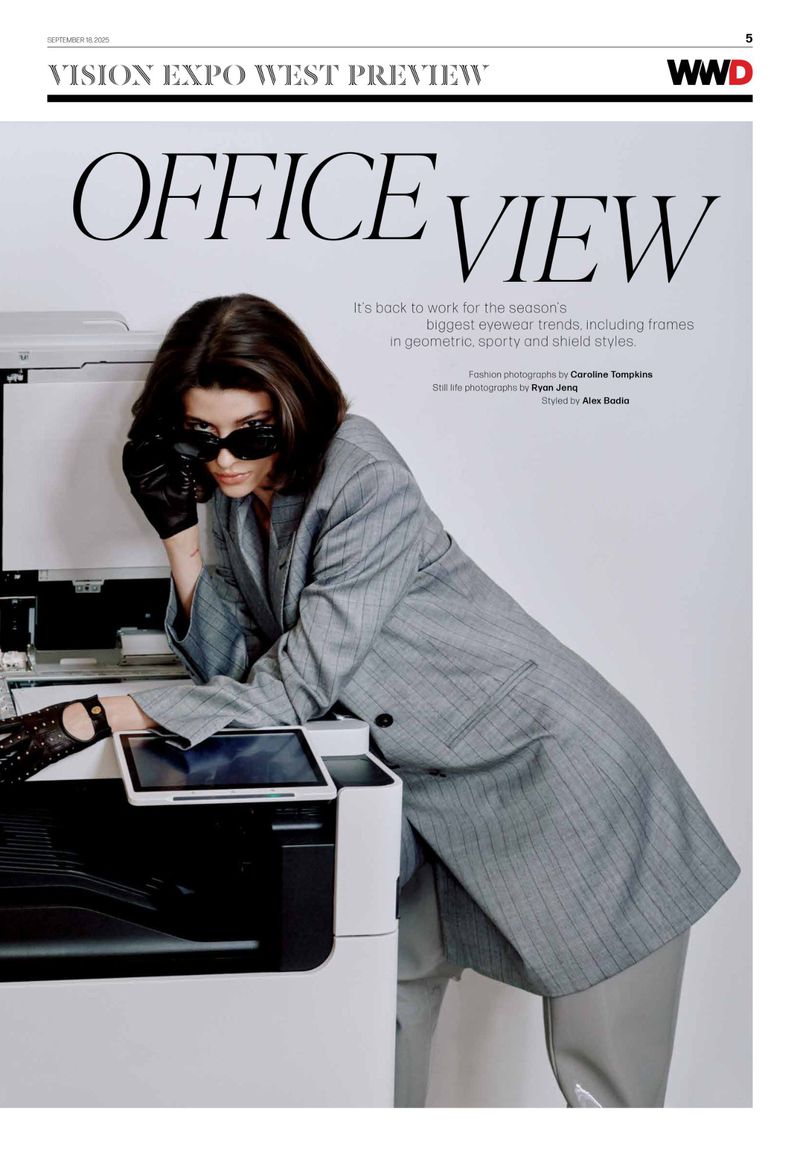 Editorial fashion shot of woman in oversized grey blazer leaning on a copier in an office, wearing dark sunglasses and black leather gloves.