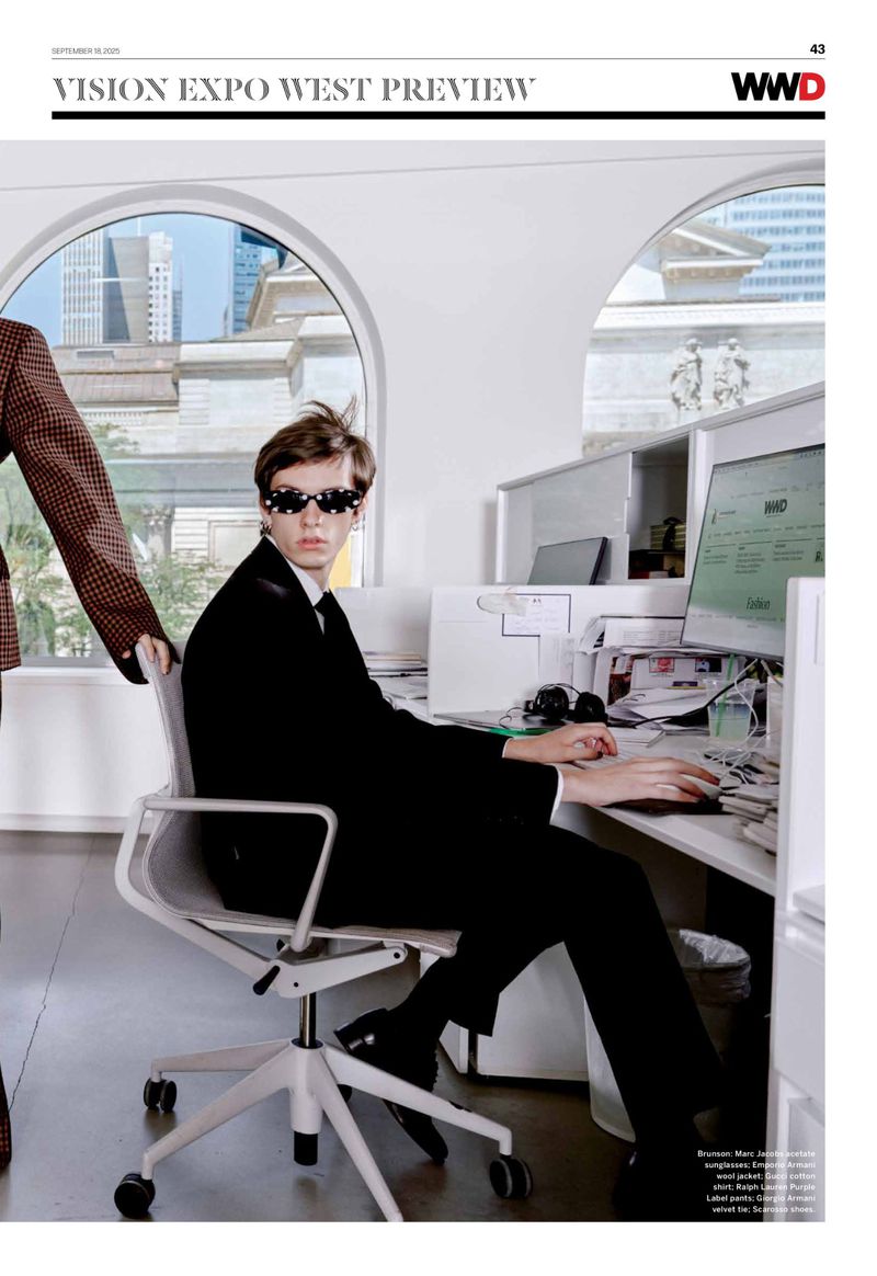 Young man in black suit and patterned sunglasses seated at a desk in office setting, with a glimpse of city skyline through windows.