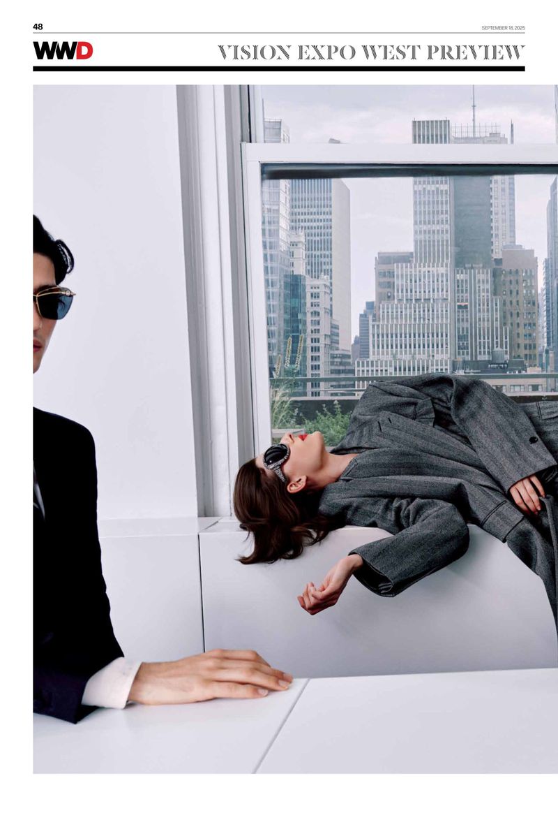 Editorial shot of two models in a studio with a cityscape view, one in a black suit and the other reclining in a gray suit.