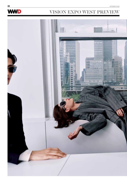 Editorial shot of two models in a studio with a cityscape view, one in a black suit and the other reclining in a gray suit.