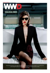 Model in dark sunglasses and black pinstripe jacket sitting on a white office desk for WWD Office View editorial