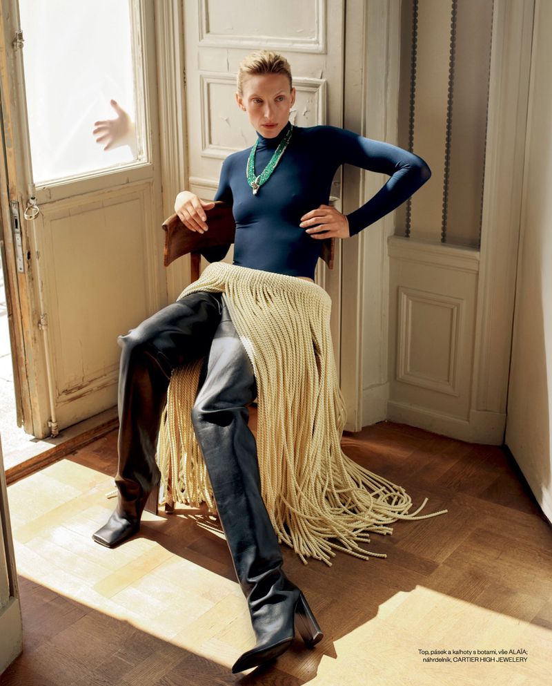 Fashion shot of a woman in a blue turtleneck, black leather thigh-high boots, and fringe seated on a wooden chair indoors.
