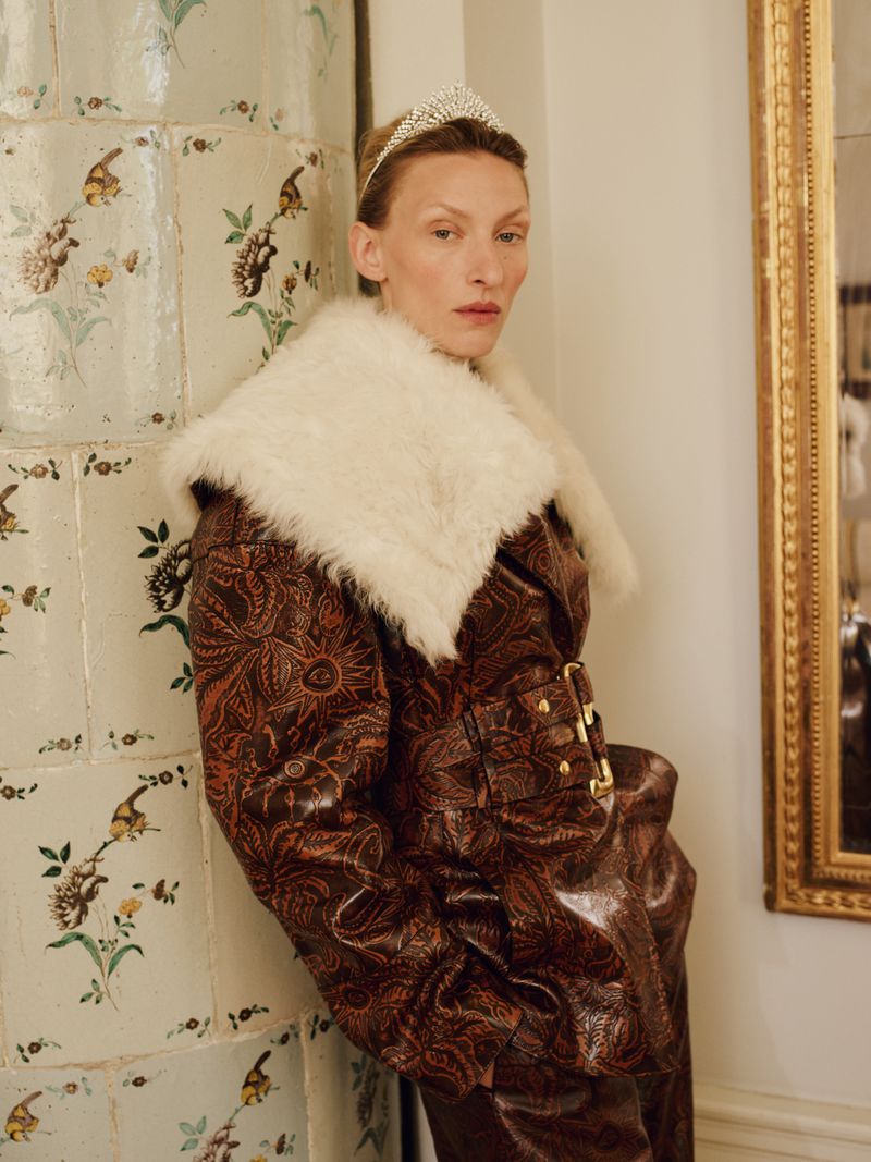 Woman in patterned jacket with fur collar and tiara posing next to floral patterned wall and gold-framed mirror in an indoor setting.
