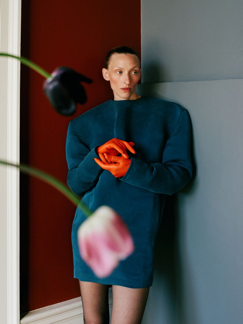 Adult female in a teal dress and orange gloves poses next to a gray and maroon wall with flowers in the foreground.