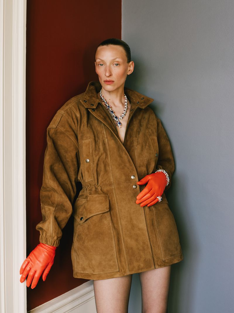 Elegant woman in a brown jacket and orange gloves poses against a corner with maroon and grey walls, showcasing fashion and style.