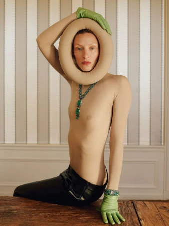Person in a beige bodysuit and black pants kneeling on a wooden table in front of a striped wall, wearing green gloves and a necklace.