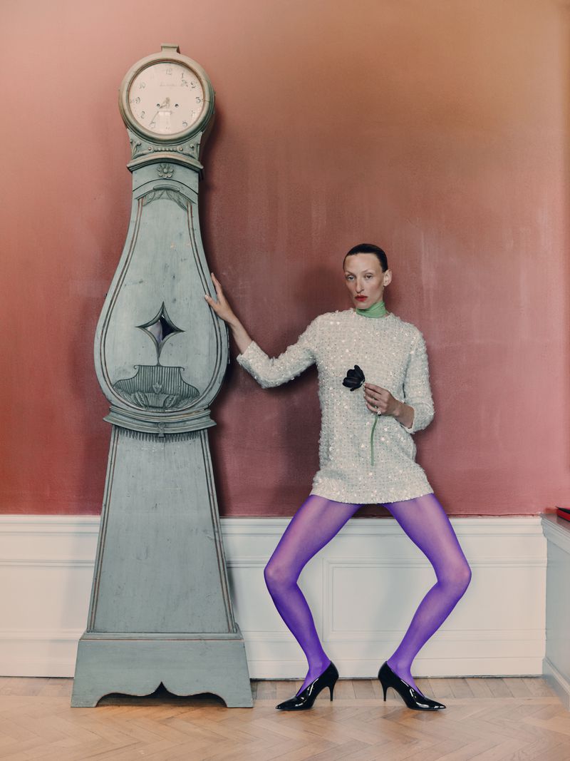 Woman poses in studio with an antique clock, wearing a sequin dress, purple tights, and heels, holding a single black rose.