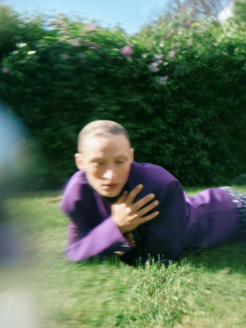 Adult lying on grass wearing a purple jacket in an outdoor garden setting, capturing a portrait with natural light and a sense of relaxation.