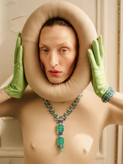 Model Maggie Maurer framed inside a circular beige padded headpiece wearing green leather gloves and an emerald statement necklace.