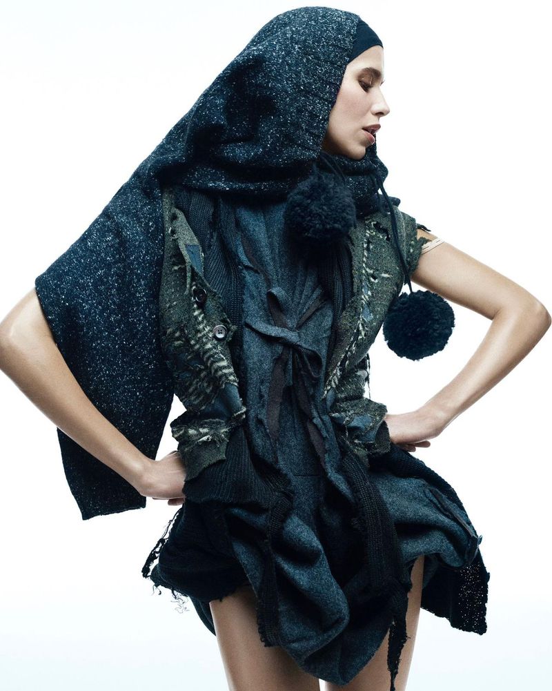 Fashion shot of a woman wearing a draped and layered outfit with a hood, featuring textile details and a dark color palette against a white background.