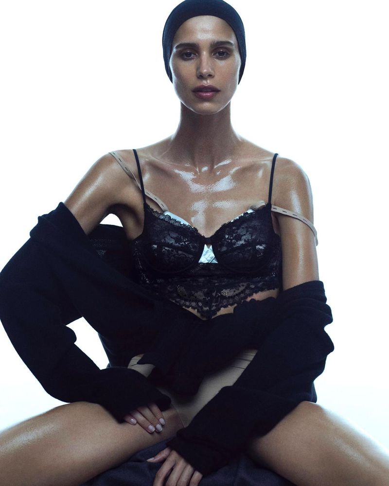 Woman in a black bralette and black wrap, seated in a bright, high-key indoor setting, presenting fashion and beauty elements.