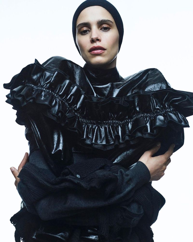Fashion portrait of a woman wearing a black head covering and dramatic ruffle-collared black top against a bright white background.