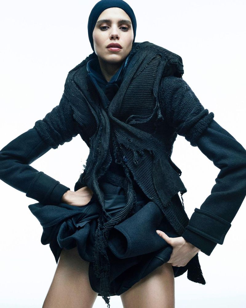 Fashionable woman in blue beanie and layered black outfit poses in a studio. Full-body shot with a bright white backdrop.