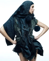 Model Mica Argañaraz posing with hands on hips wearing layered dark knitwear and a hooded scarf against a white studio background.