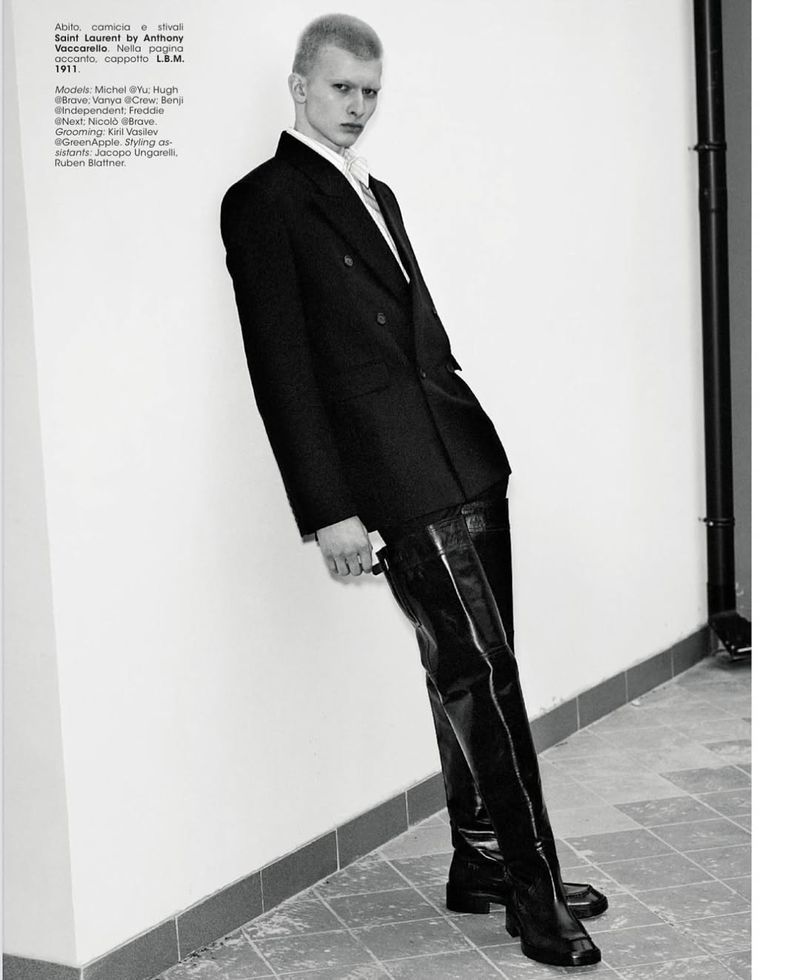 A male fashion model with short hair poses in a black blazer and shiny thigh-high boots against a neutral wall and tiled floor.