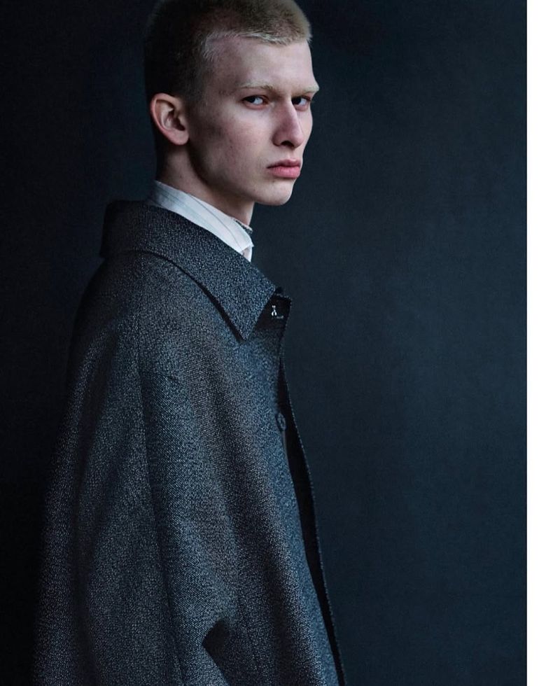 A young man with a blonde buzz cut wearing a textured grey overcoat and looking over his shoulder against a dark background.
