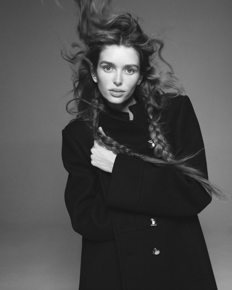 Monochrome portrait of a woman with long, flowing hair styled in braids, wearing a black coat against a neutral grey backdrop in studio.