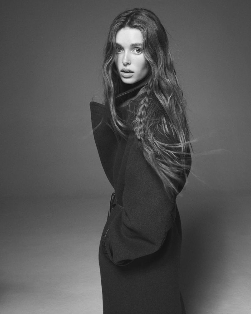 Monochrome portrait of an adult woman with long, wavy hair wearing a dark, belted coat in a photo studio against a grey backdrop.