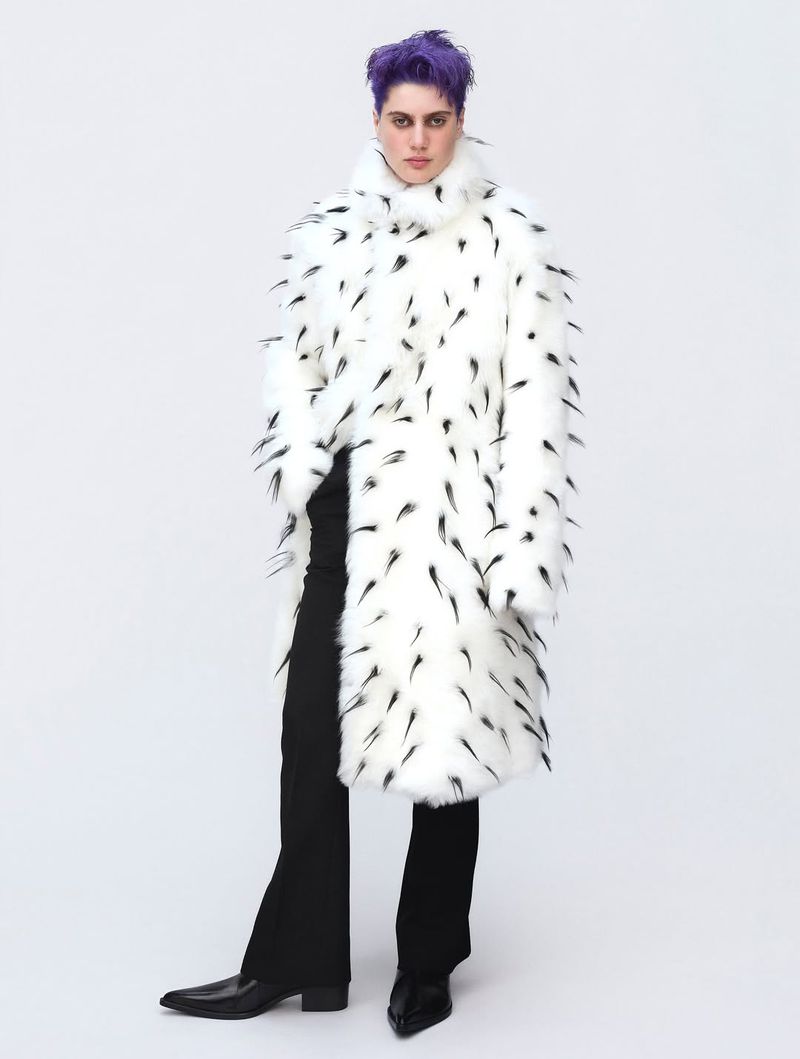 Person with purple hair stands in a bright studio wearing a long white faux-fur coat with black markings and black pants and shiny shoes.