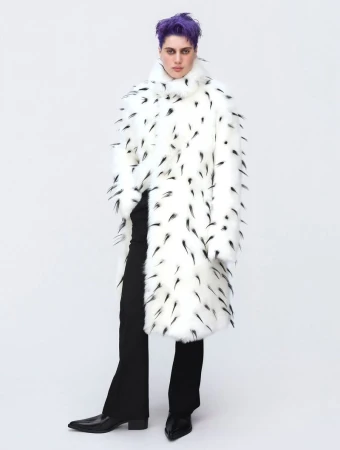 Person with purple hair stands in a bright studio wearing a long white faux-fur coat with black markings and black pants and shiny shoes.